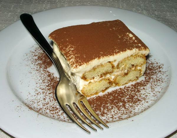A Slice of tiramisu cake on a white plate with chocolate powder sprinkled over the top.