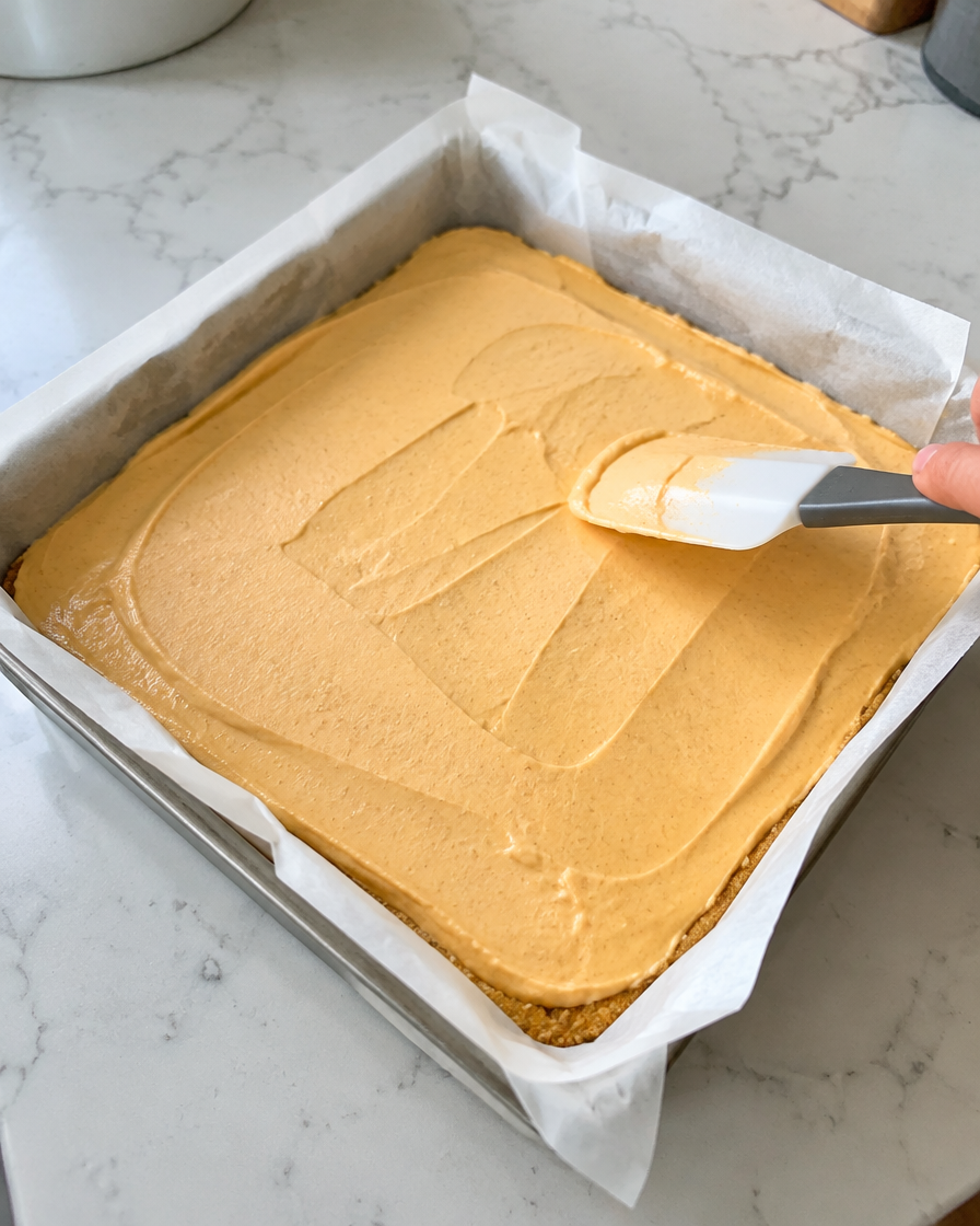 Spreading pumpkin cheesecake filling over the crust in a parchment-lined square pan